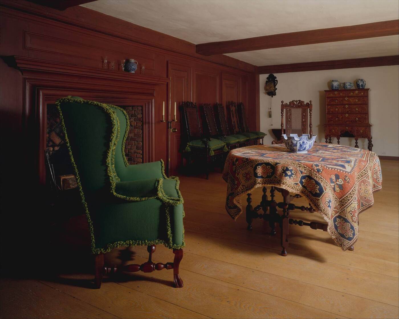 A paneled colonial room with a green wing chair and a gateleg table draped in an antique carpet